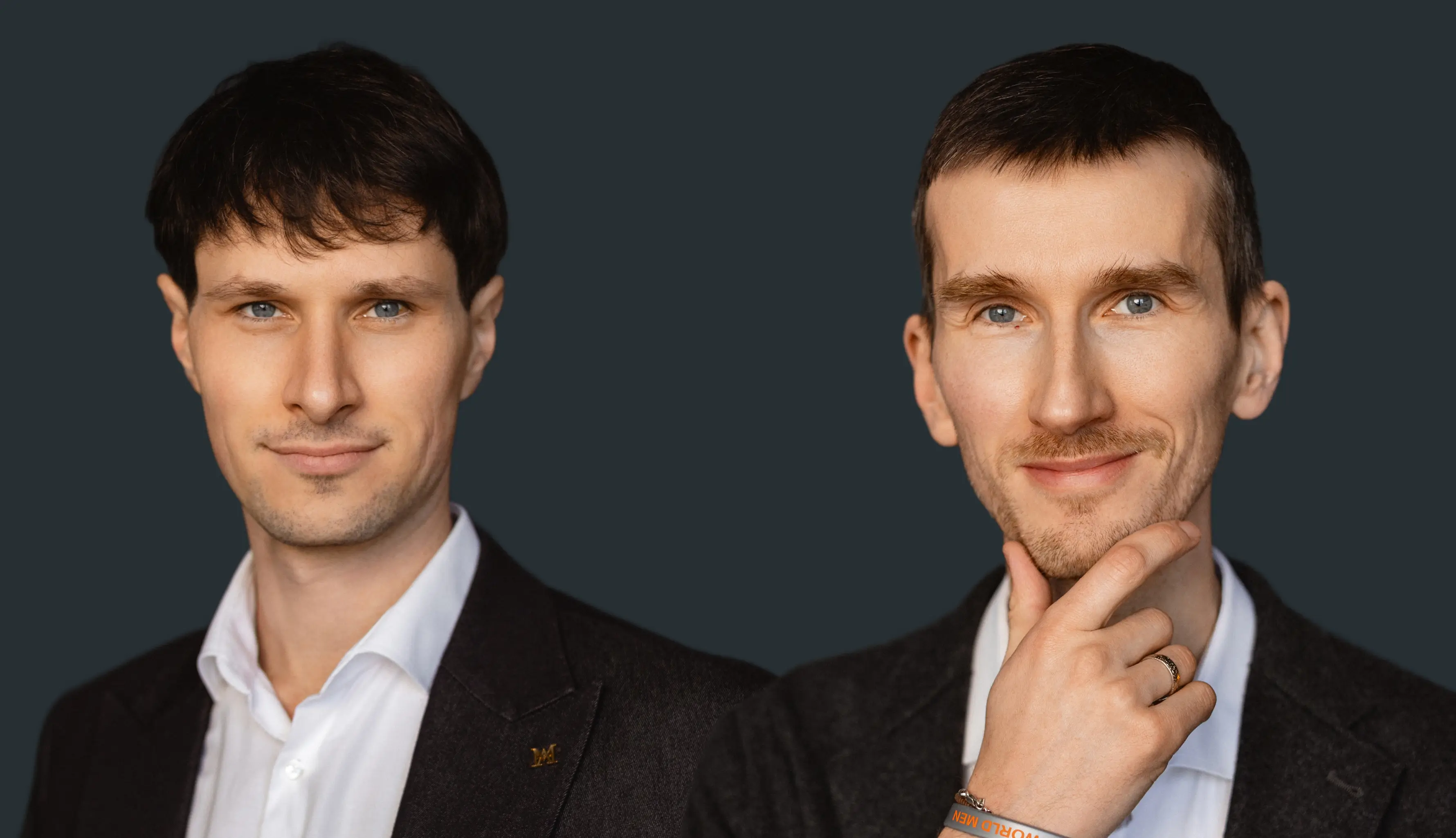 Two men in dark blazers and white shirts against a dark background, one smiling with hand on chin.