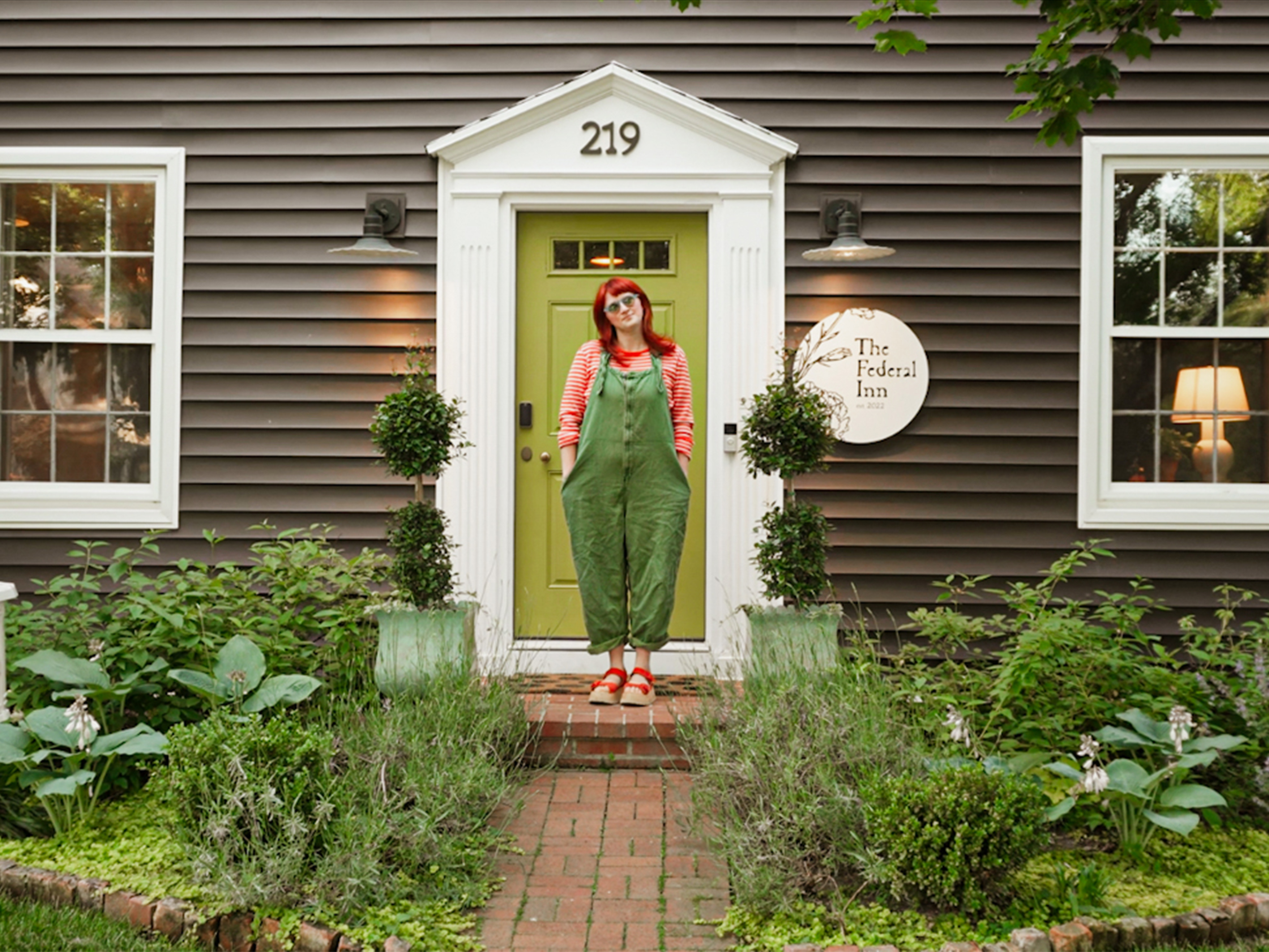 A person with red hair, sunglasses, red striped shirt, and green overalls stands with hands in pockets in front of a green door at 219 of a brown siding building with white trim and two windows, one with a lit lamp. Two potted topiary plants flank the door, and a round sign reading 'The Federal Inn est 2022' is on the wall.
