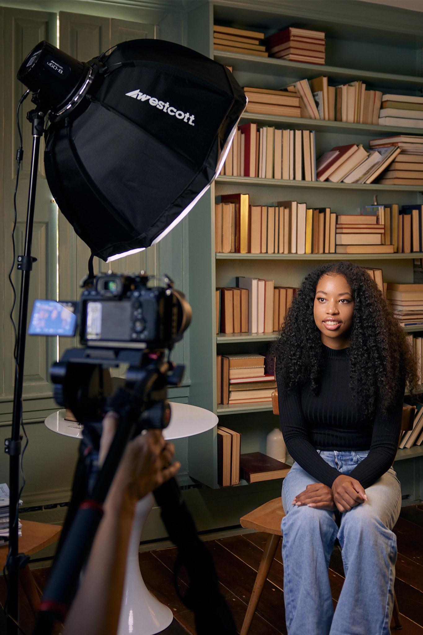 Young woman with curly hair wearing a black long-sleeve top and light blue jeans sits on a wooden chair in front of a bookshelf filled with books, being recorded by a camera with a large Westcott softbox light in a cozy indoor setting.