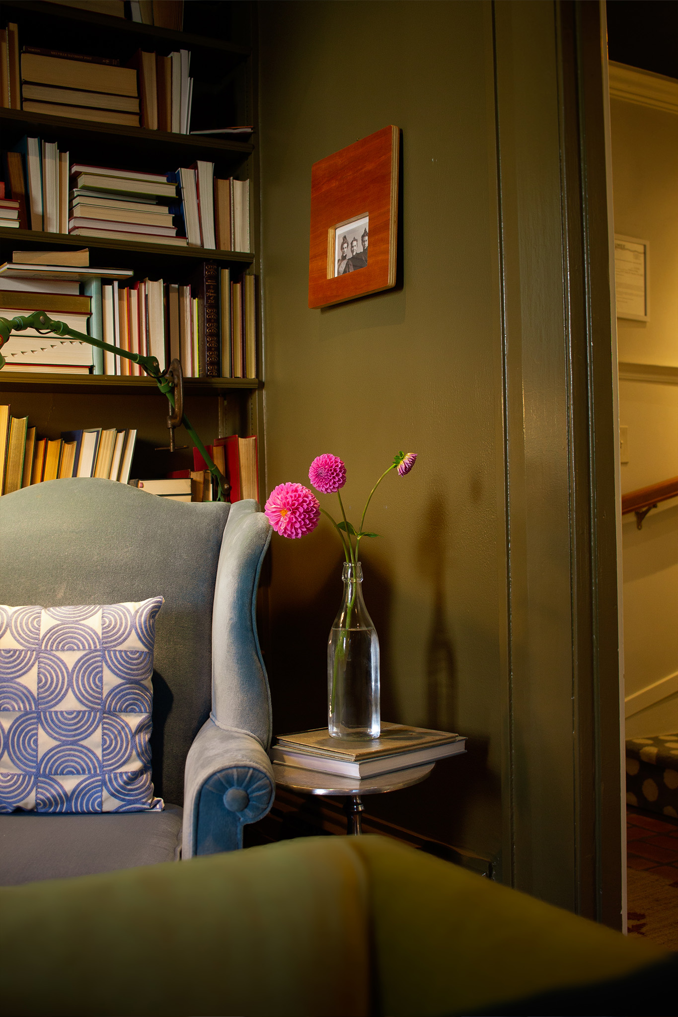 Cozy corner with a blue upholstered armchair featuring a blue and white geometric patterned pillow, a small round table holding a glass bottle with water and three pink flowers, and a bookshelf filled with books in the background, framed by olive green walls and a small wooden picture frame with a black and white photo.