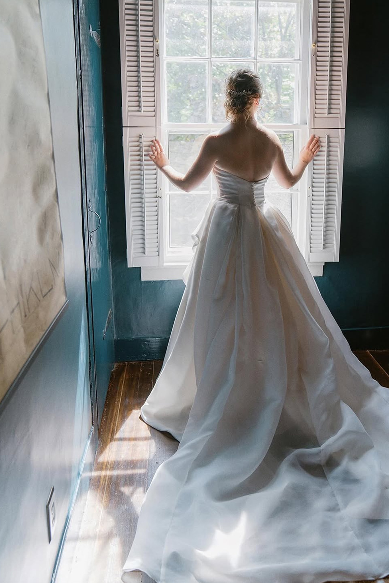 Bride in a strapless white wedding gown standing with her back to the camera, opening window shutters to let sunlight into a room with dark teal walls and wooden floors.