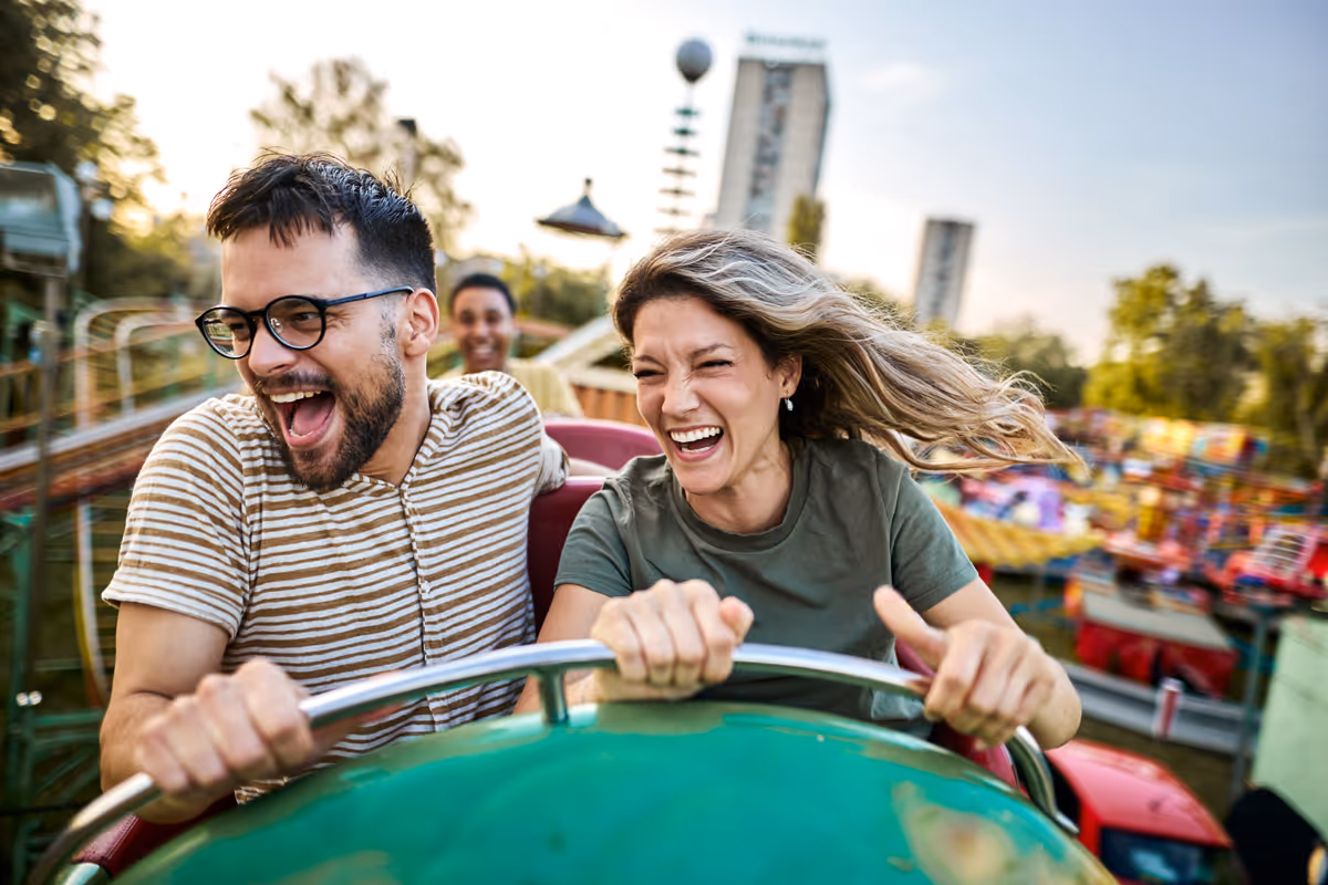 Two young adults laughing and holding onto safety bar while riding a roller coaster at an amusement park.