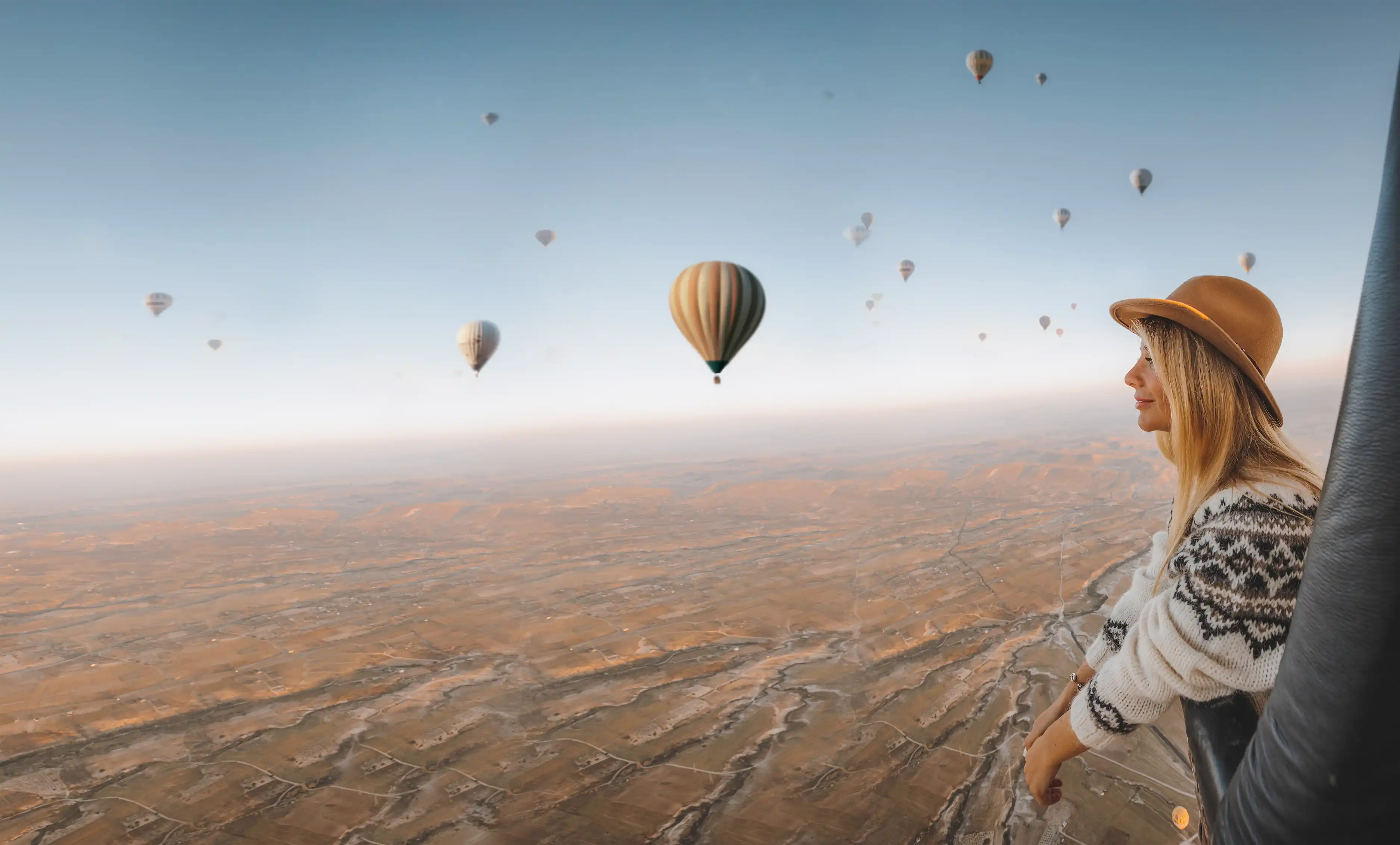 Woman wearing a patterned sweater and brown hat looking out from a hot air balloon at multiple hot air balloons over a vast landscape.