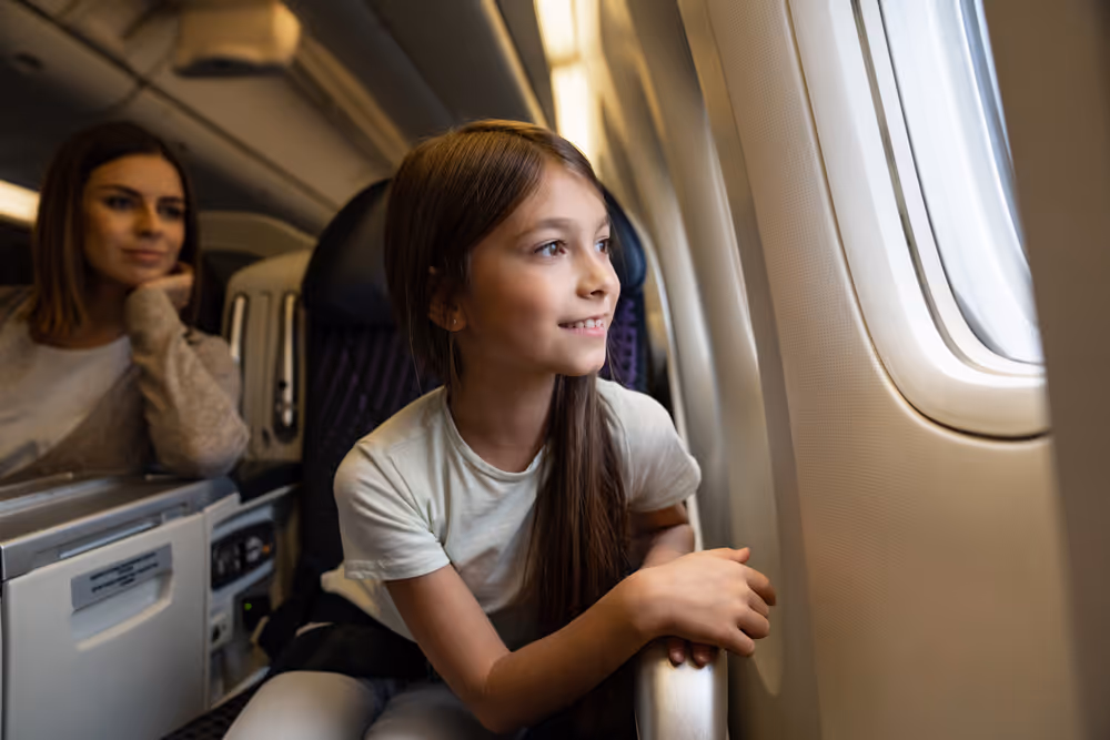 Young girl with long brown hair looking out the window of an airplane while a woman sits behind her.