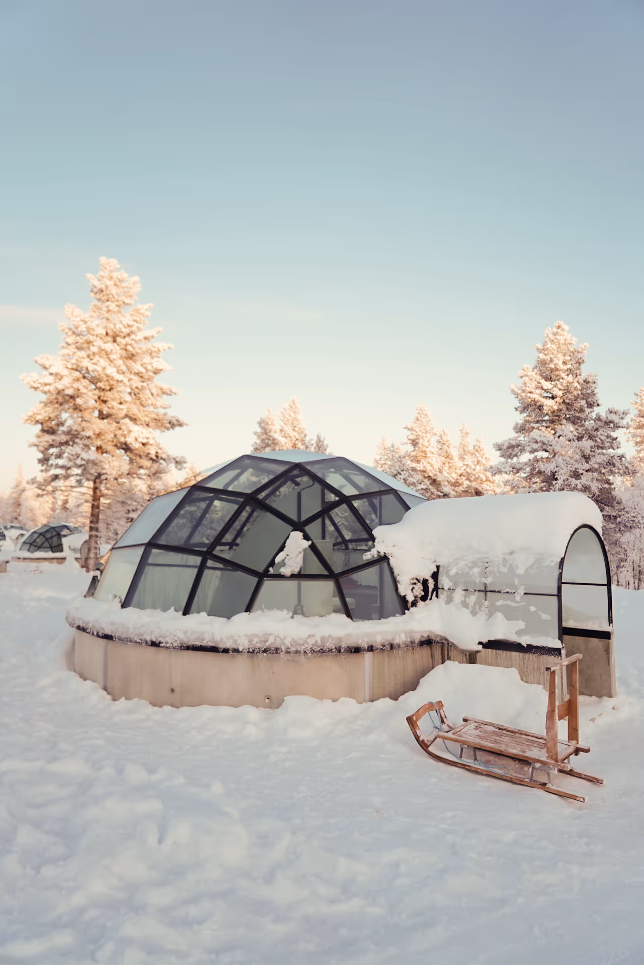 Glass-domed igloo cabin covered in snow with a wooden sled in front, set in a winter landscape with snow-covered trees under a clear sky.