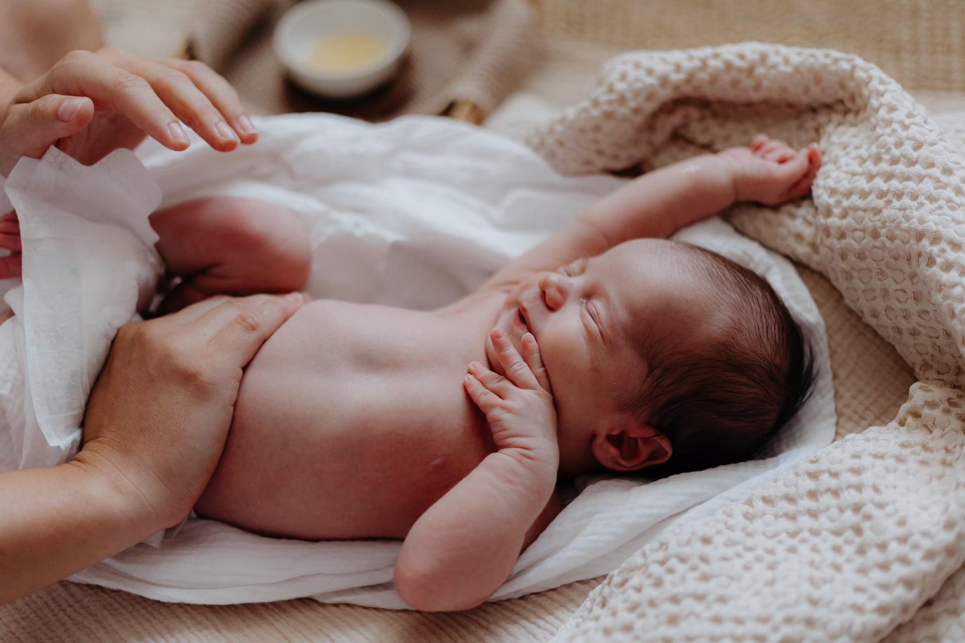 Newborn baby lying on a blanket with a hand gently holding its side, smiling with eyes closed.