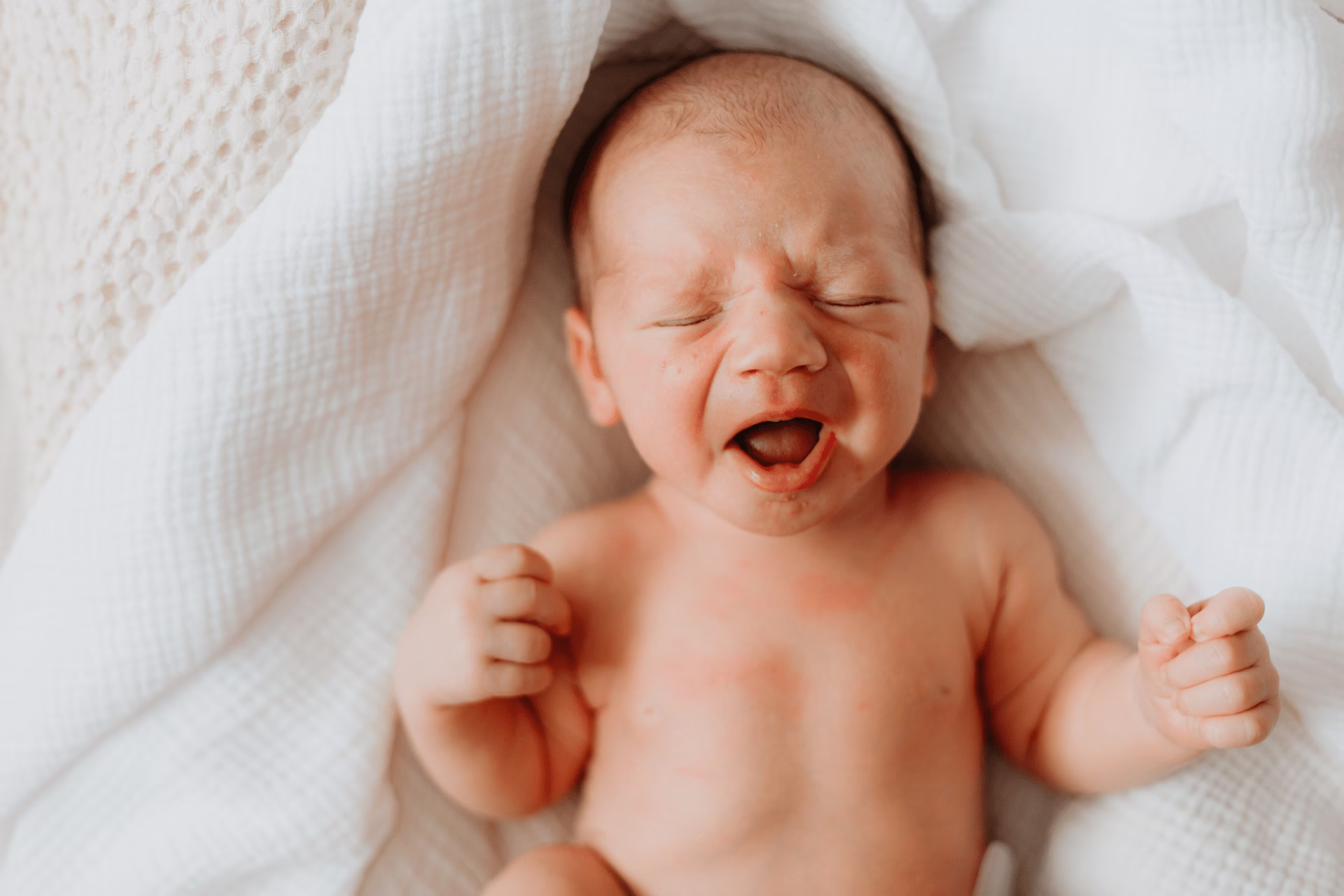 Newborn baby wrapped in white blankets with eyes closed and mouth open, appearing to cry.