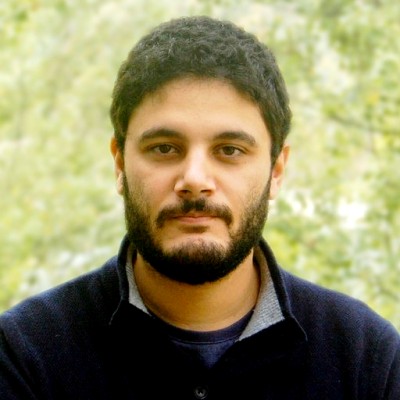 Portrait of a man with short curly dark hair, full beard, and a serious expression wearing a dark jacket, with a blurred green background.