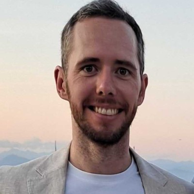 Smiling man with short hair and beard wearing a light gray blazer and white shirt against a backdrop of mountains at sunset.