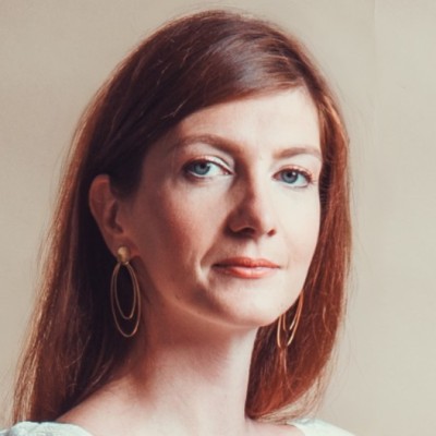 Portrait of a woman with long auburn hair, wearing gold hoop earrings and light makeup, looking slightly to the side.