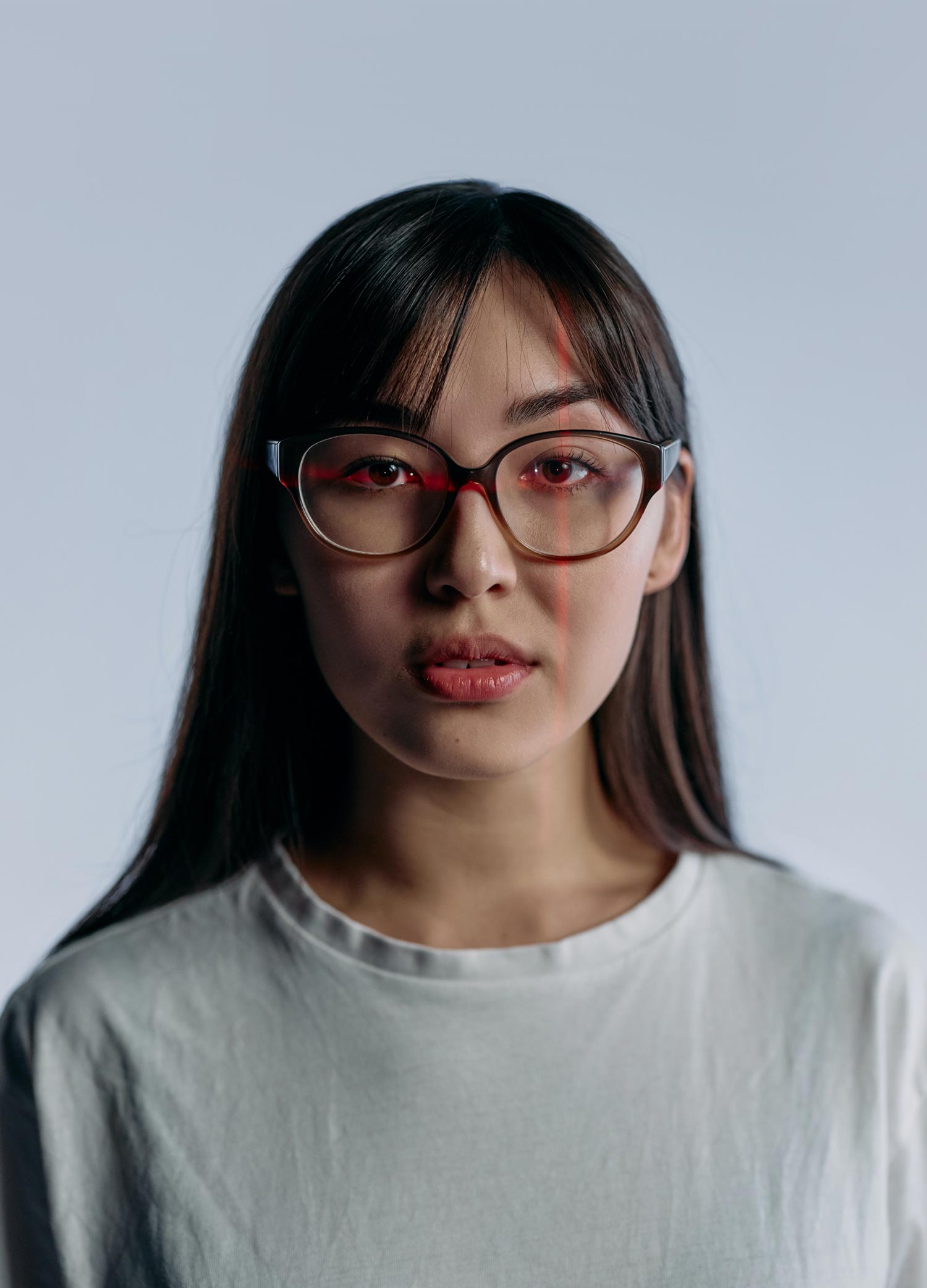 Young woman with long dark hair and red-rimmed glasses, wearing a light T-shirt against a plain pale background, with faint red laser lines intersecting over her face.
