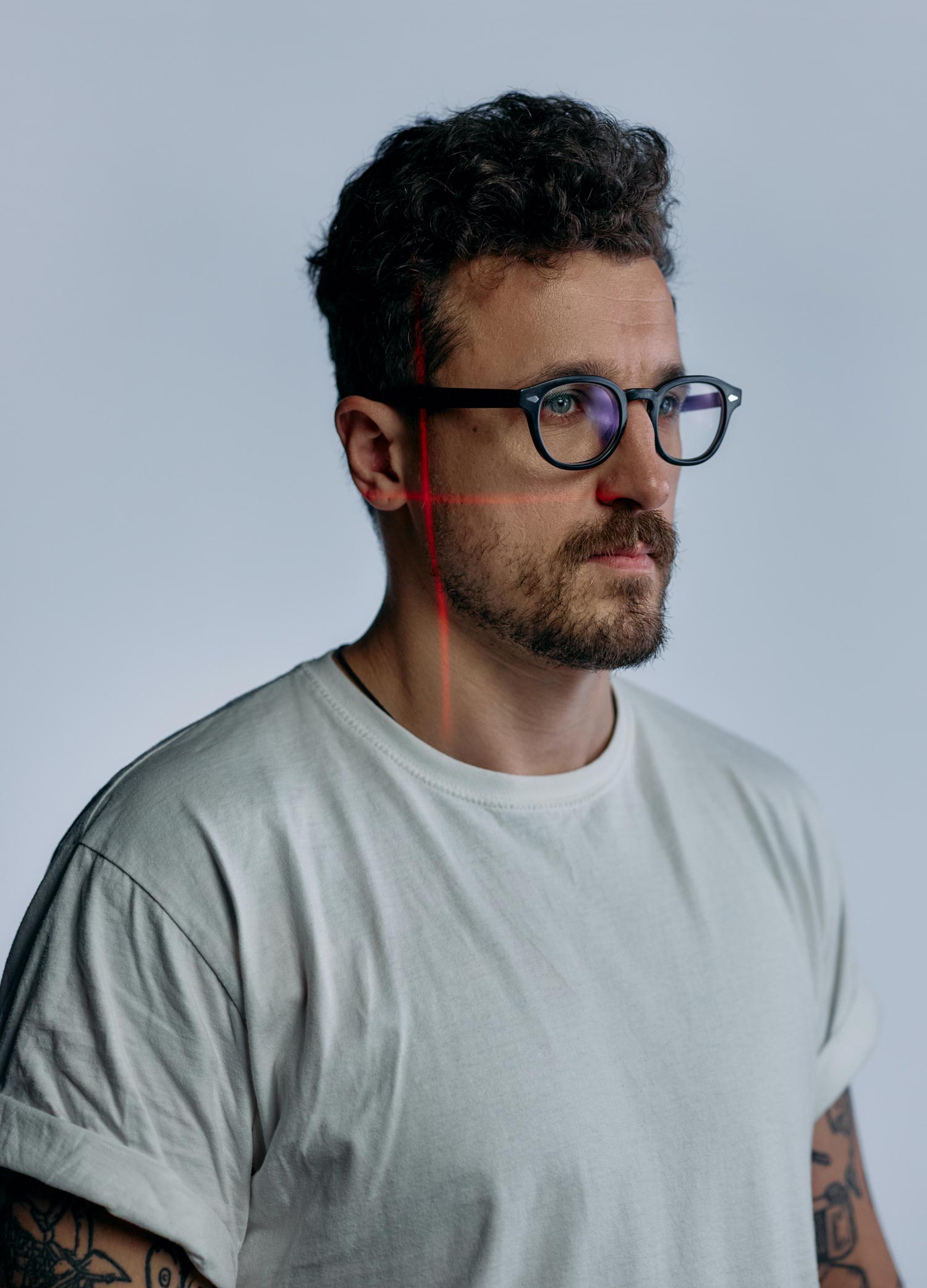 Bearded man with glasses and tattooed arms wearing a light T-shirt, seen in profile against a plain background while thin red laser lines form a vertical and horizontal cross on his face.