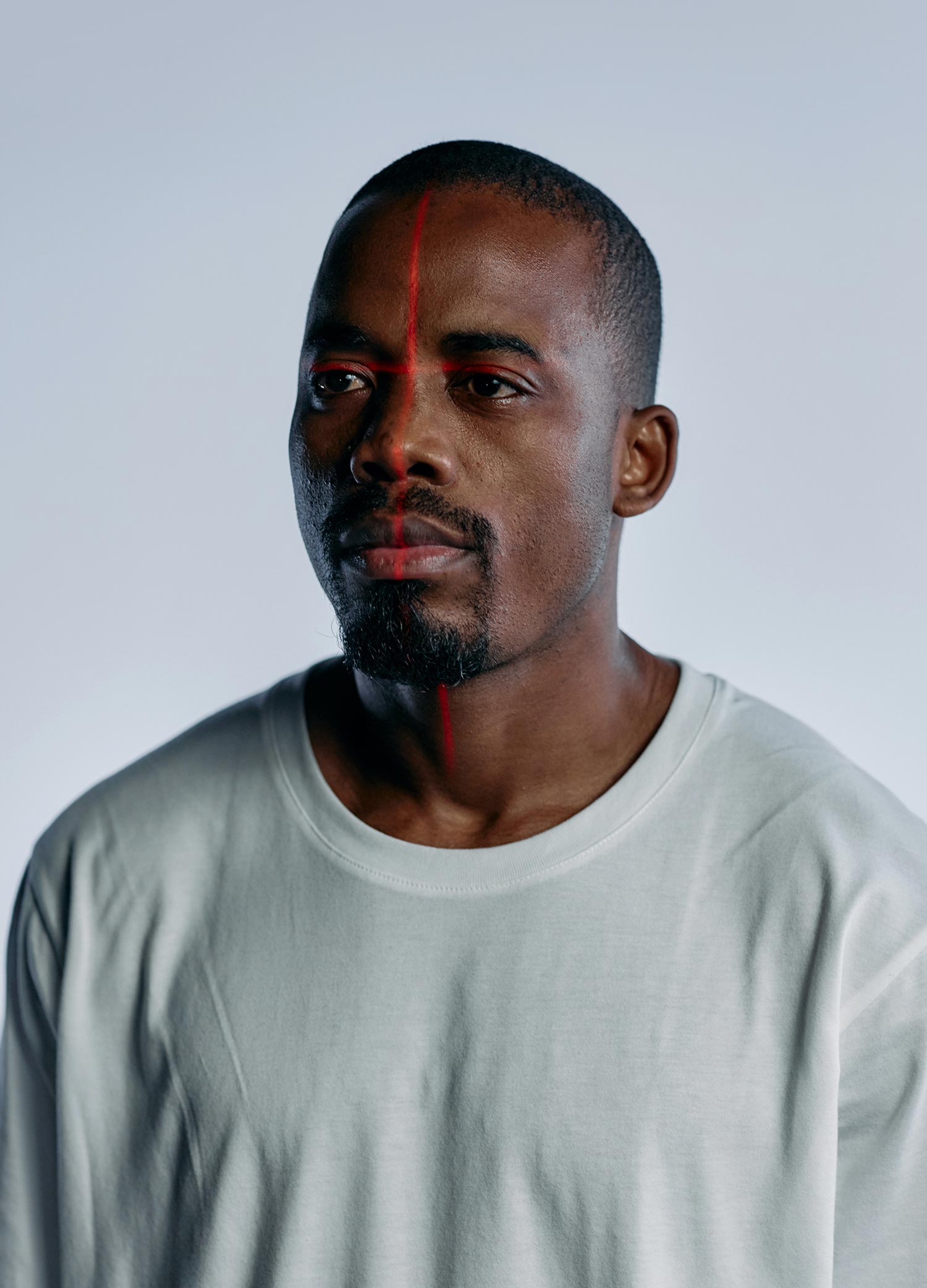 Portrait of a Black man in a light gray T-shirt against a plain pale background, with a thin vertical red laser line running down the center of his face.