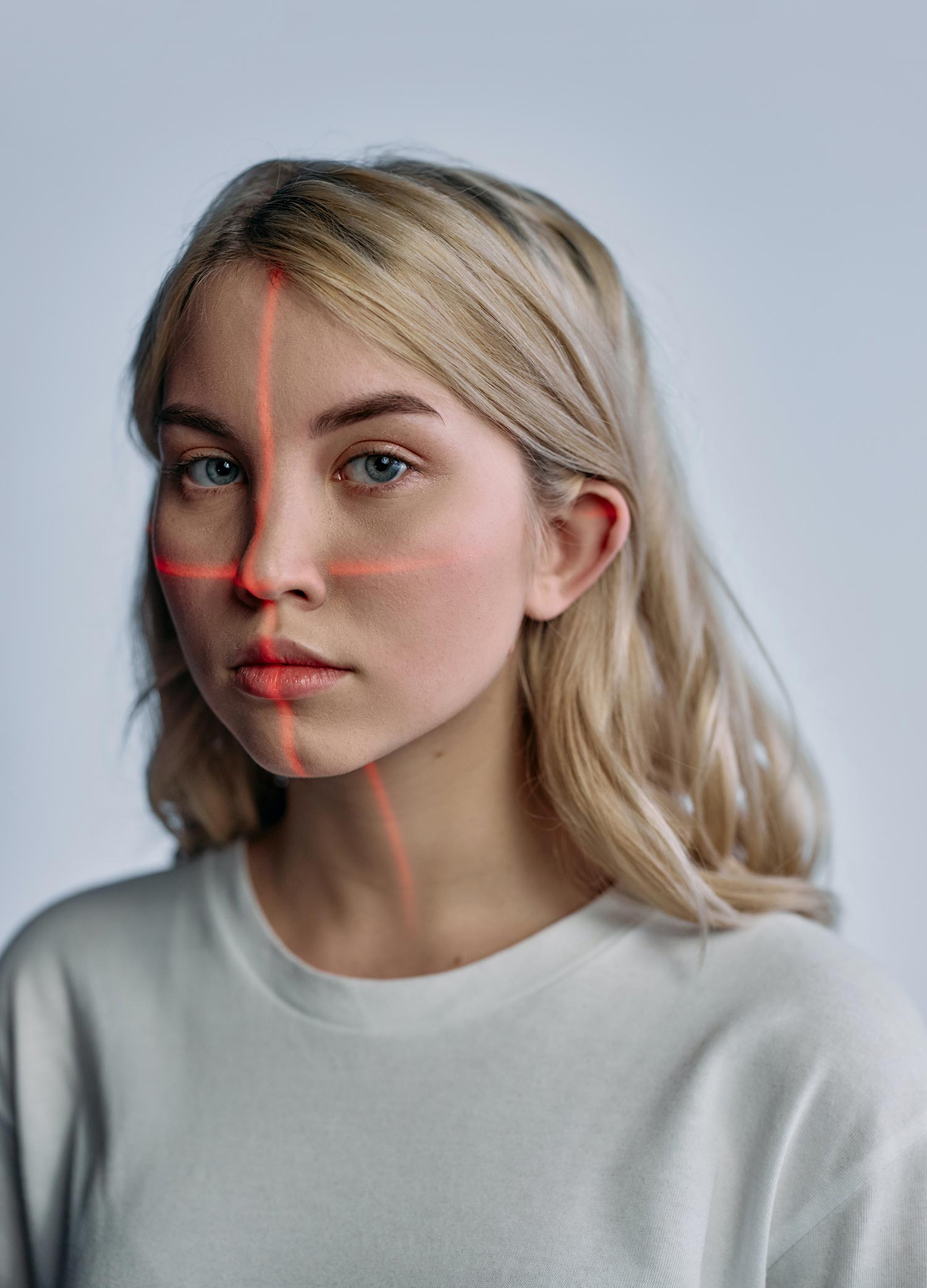 Portrait of a young blonde woman in a light crew-neck shirt, facing forward while red laser lines form a vertical and horizontal cross over her face against a plain pale background.