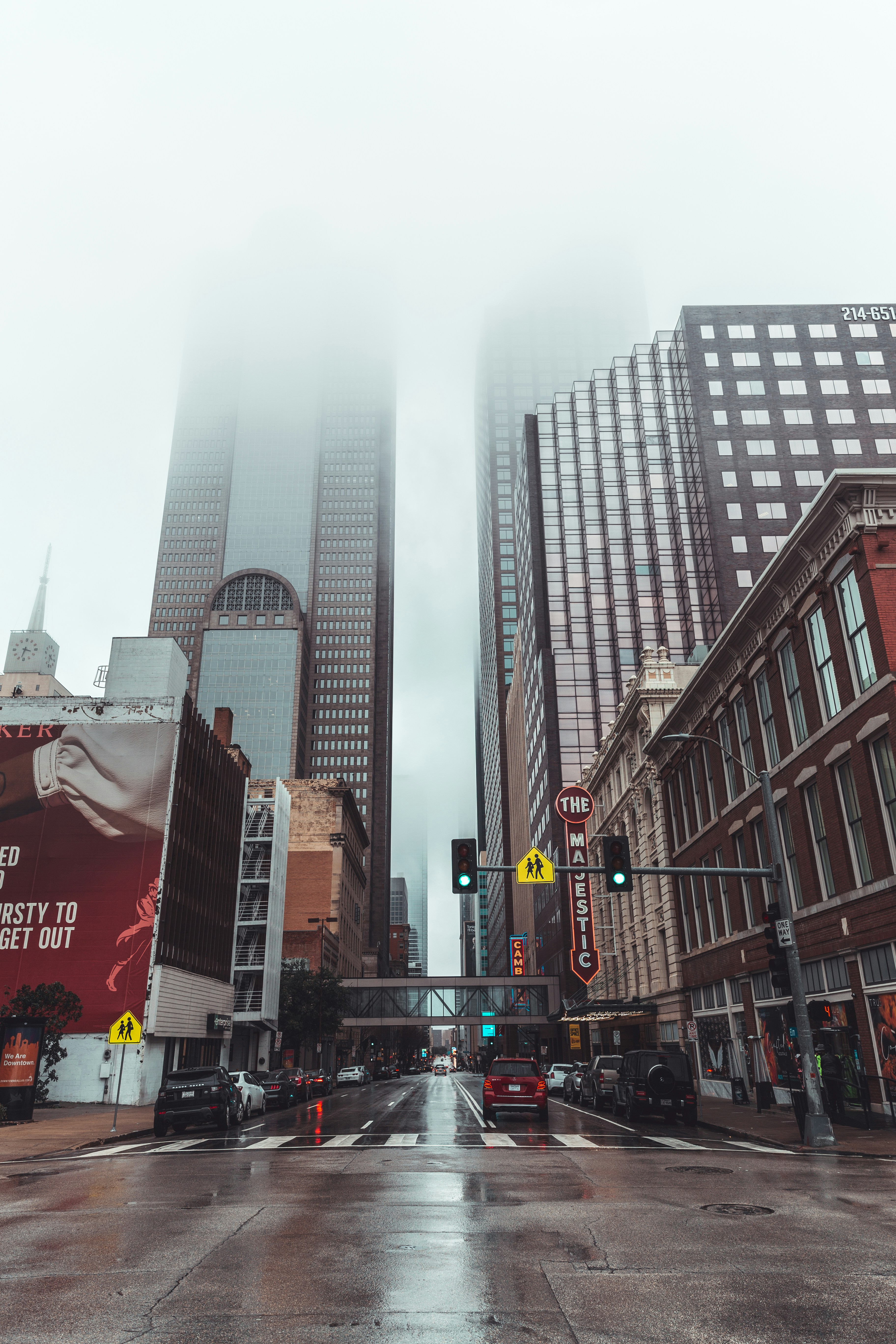 Foggy downtown Dallas street view with tall skyscrapers disappearing into low-hanging clouds, featuring the historic Majestic Theatre sign, wet pavement from recent rain, and cars stopped at a green pedestrian crossing light.