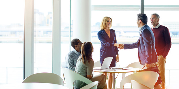 Business professionals in a bright office meeting space, with a woman shaking hands with a man while two colleagues look on, seated at a table with a laptop near large windows.