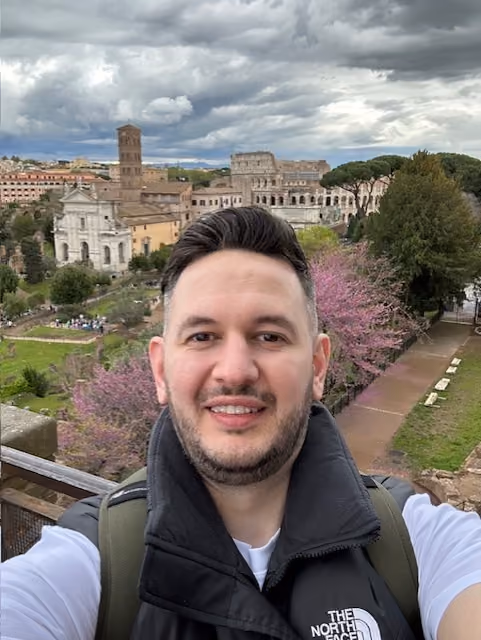 Man with dark hair and beard wearing a black North Face vest and white shirt smiling in front of the Roman Colosseum and ancient buildings under a cloudy sky with trees and pathways nearby.