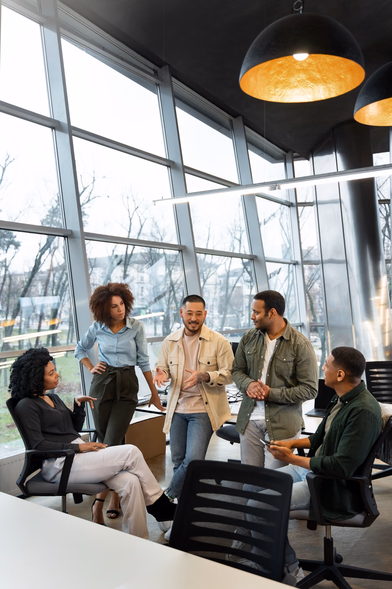 Five young professionals engaged in a discussion in a modern office with large glass windows and black hanging lamps, with one man gesturing while talking and others attentively listening.