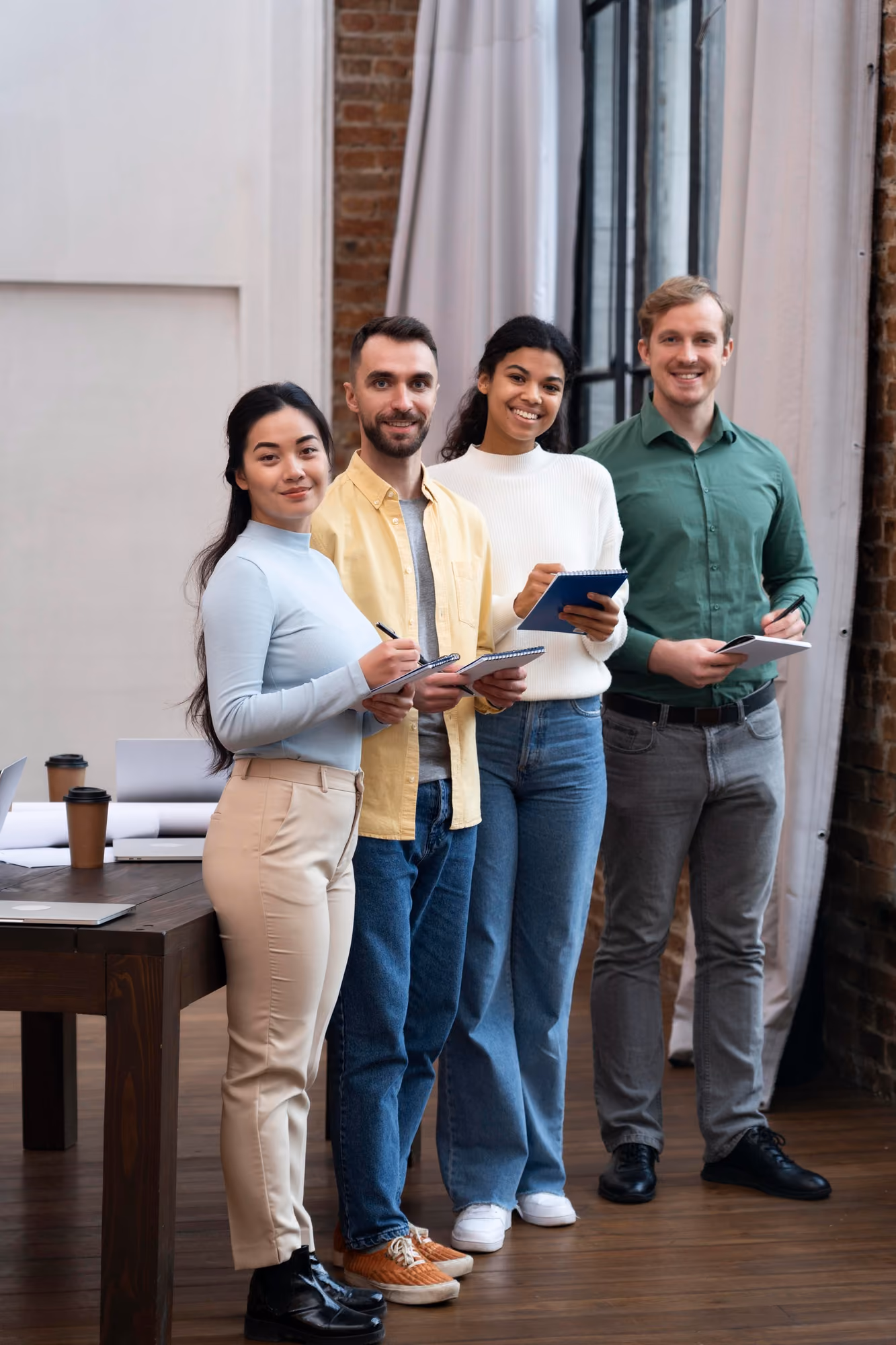 Four diverse coworkers standing side by side in a modern office, holding notebooks and pens, smiling at the camera.