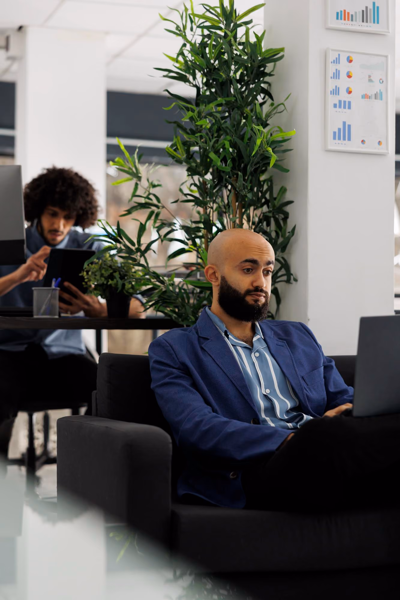 Man with a beard and bald head wearing a blue blazer and striped shirt, sitting on a black couch working on a laptop, with another man in the background using a tablet at a desk beside green plants in a modern office.