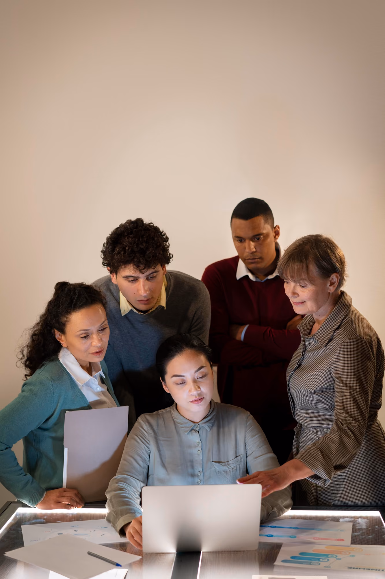 Five coworkers gathered around a laptop, focused on the screen, collaborating on a project with printed charts on the table.
