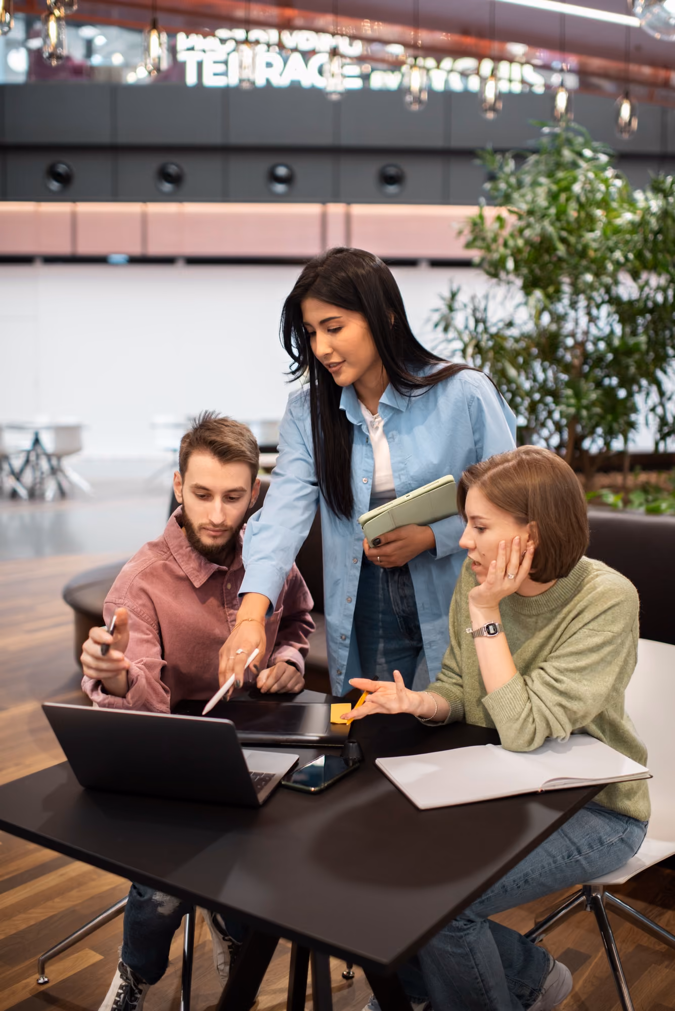 Three young adults collaborating around a laptop at a black table in a modern indoor space, with one woman standing and pointing at the screen while holding a notebook, and a man and another woman seated, engaged in discussion.