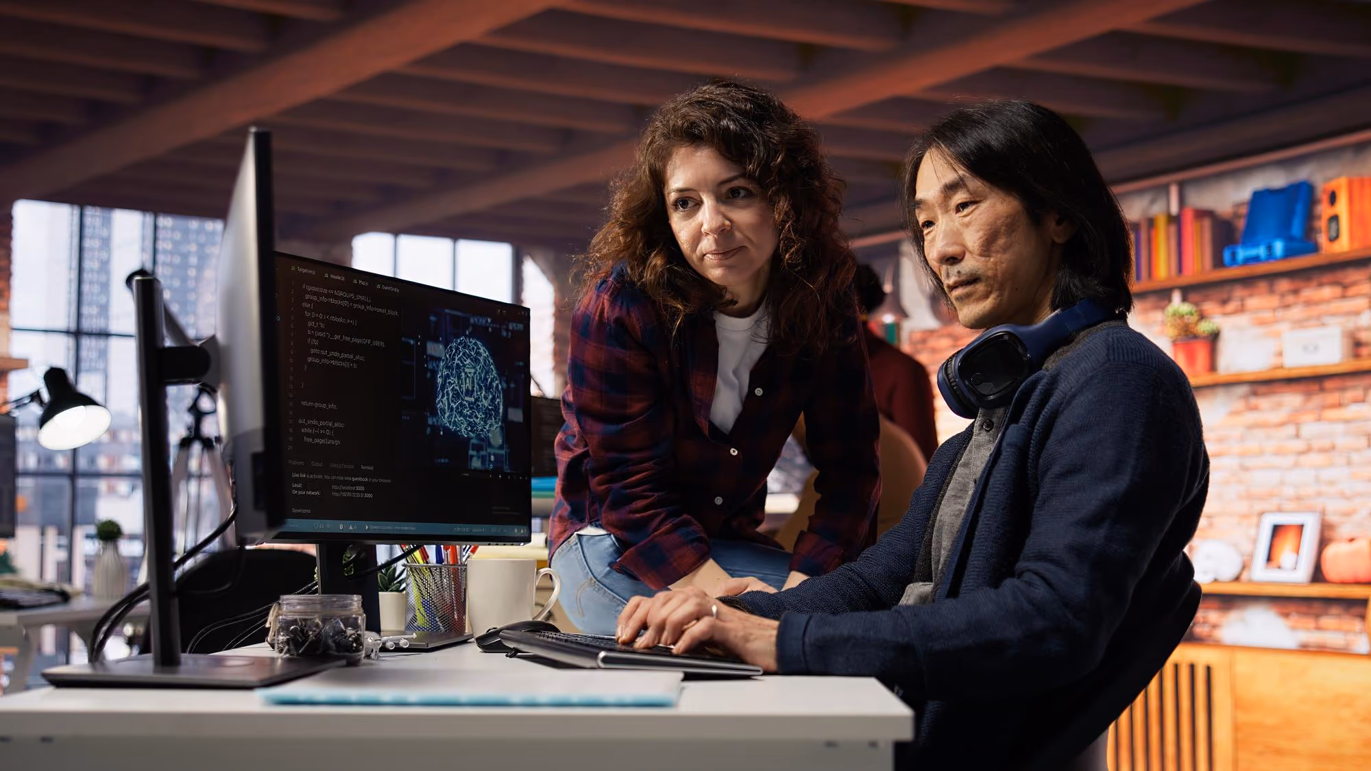 Two colleagues in a modern office working together at a computer displaying code and a brain graphic, with one person seated and typing and the other leaning in and observing.