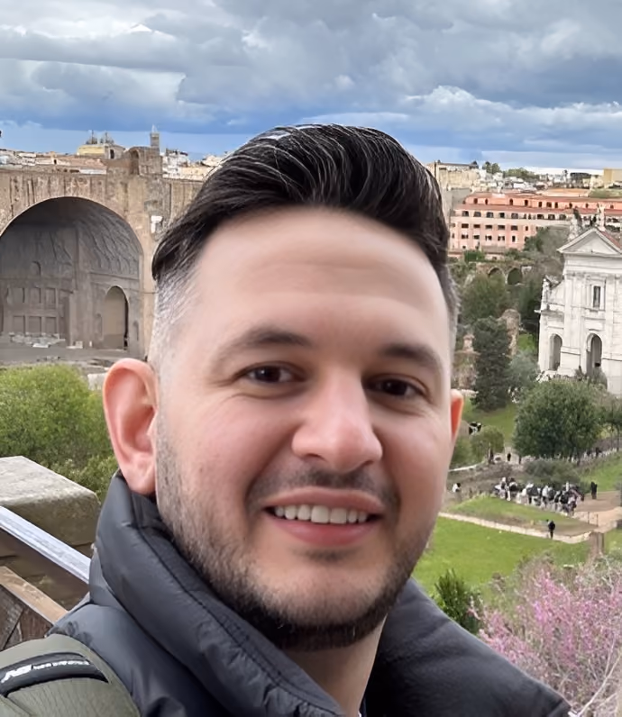 Close-up of a smiling man with short dark hair and a trimmed beard wearing a black jacket outdoors with historic ruins and greenery in the background under a cloudy sky.