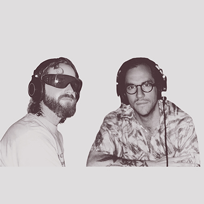 Two men from group Two Planets wearing headphones; one has a beard and large sunglasses, the other wears glasses and a patterned shirt.