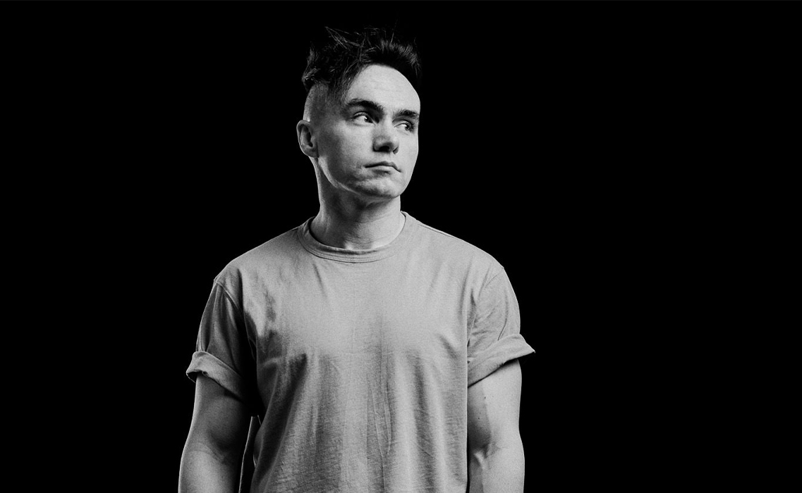 Black and white portrait of Producer and writer Lewis Gardiner with short, styled hair wearing a rolled-up sleeve t-shirt looking to the side against a black background.
