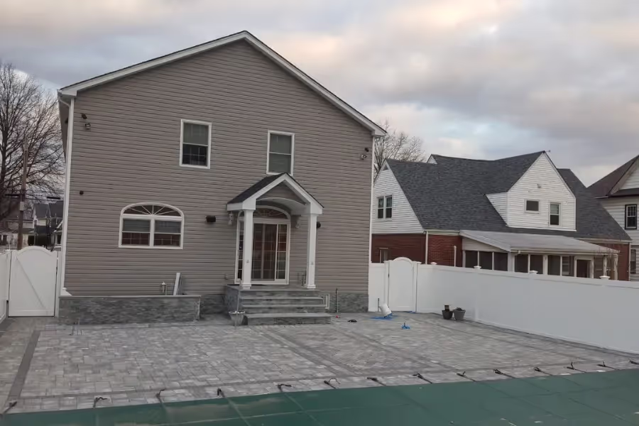 Backyard of a two-story house with gray siding and a covered porch, featuring a large paved patio area and white privacy fences.