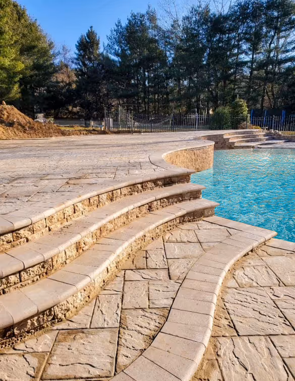 Curved stone steps leading down to a swimming pool with clear blue water, surrounded by patterned stone paving and trees in the background.