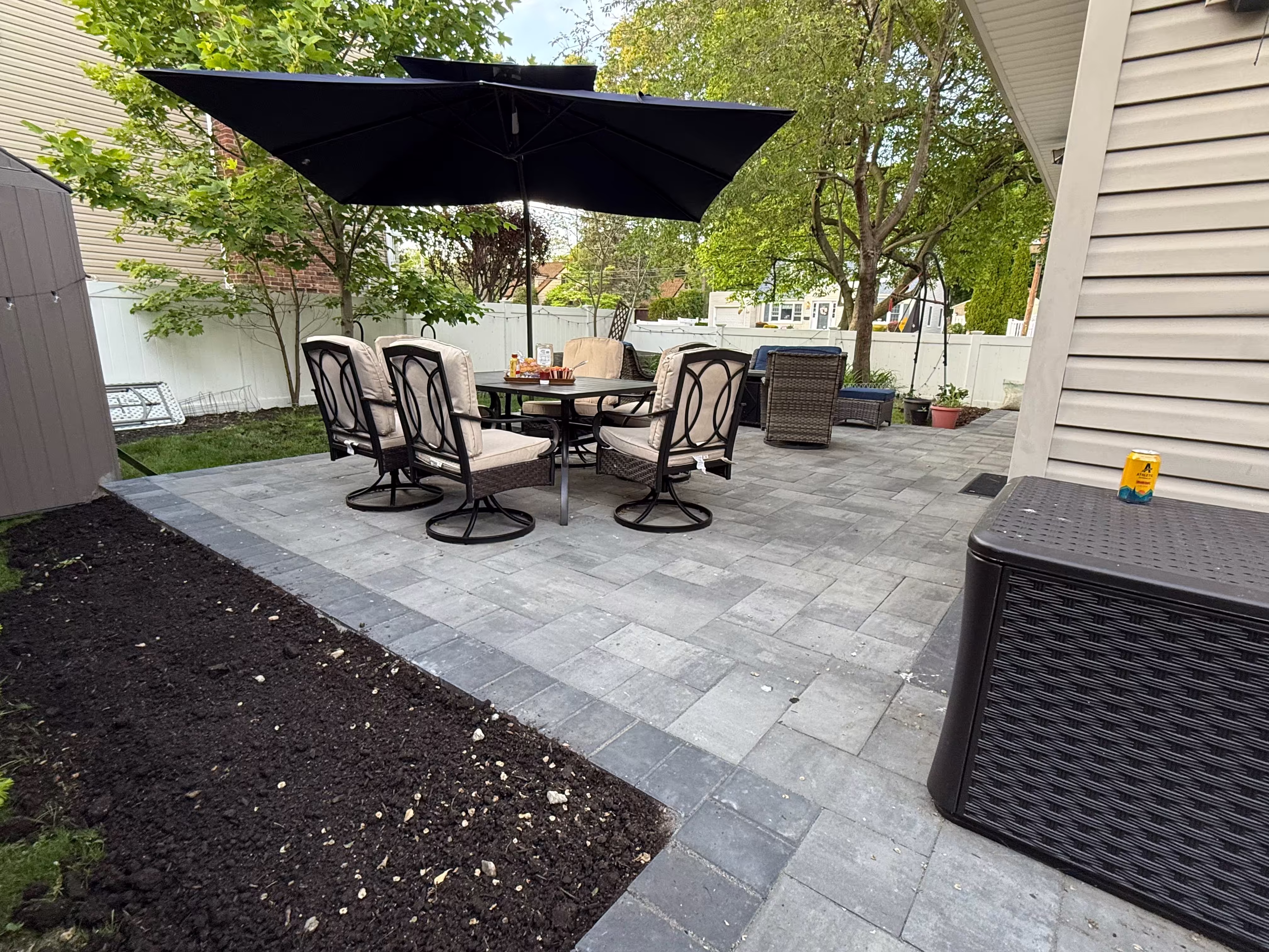 Backyard patio with gray stone tiles, outdoor dining table with six cushioned chairs under a large black umbrella, and surrounding greenery.