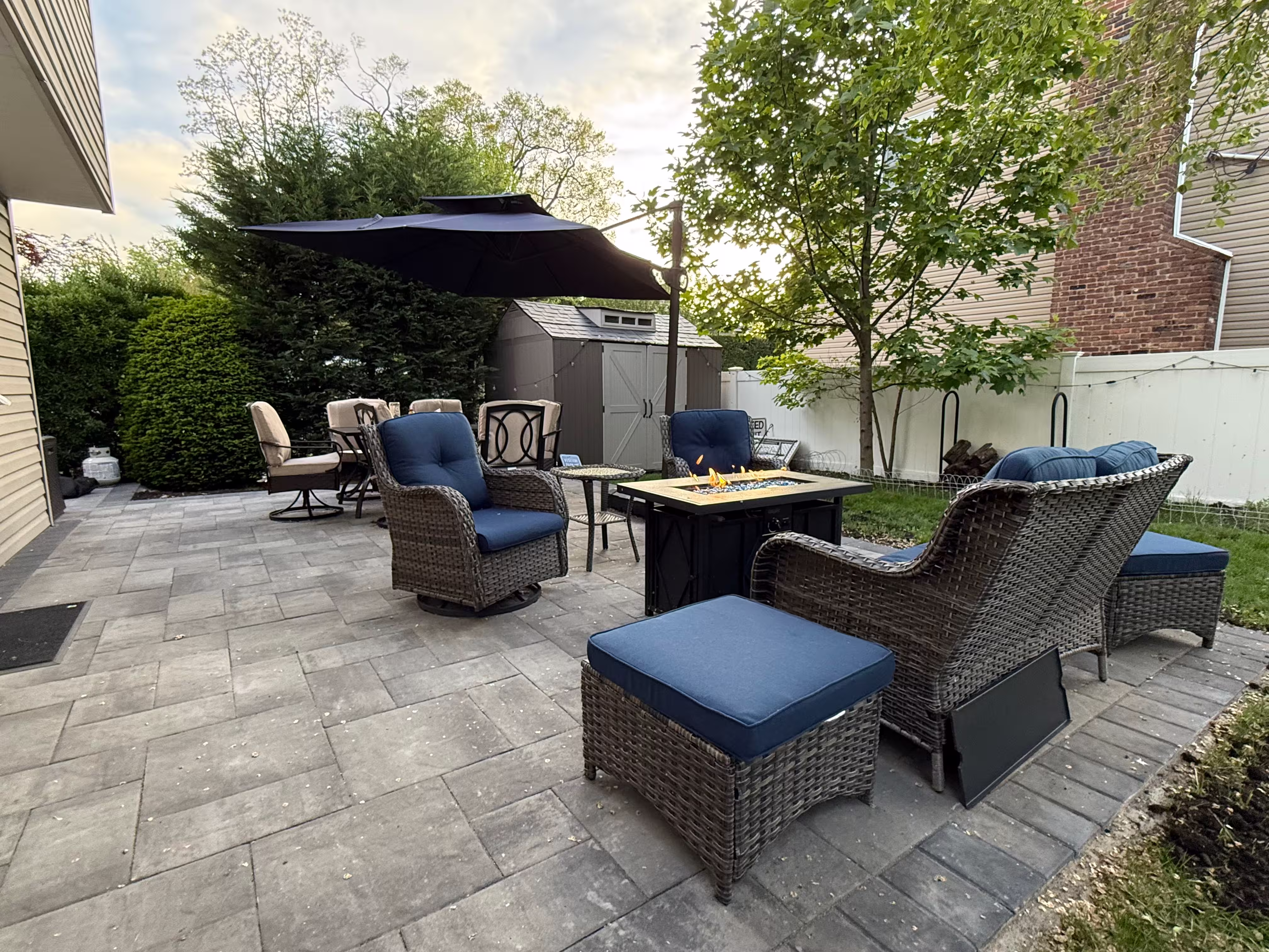 Outdoor patio with grey stone tiles, wicker chairs with blue cushions around a fire pit, a dining table with beige chairs under a large dark umbrella, and a brown storage shed near a white fence.