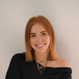 Smiling woman with red hair wearing a black off-shoulder top and layered gold necklaces against a light background.