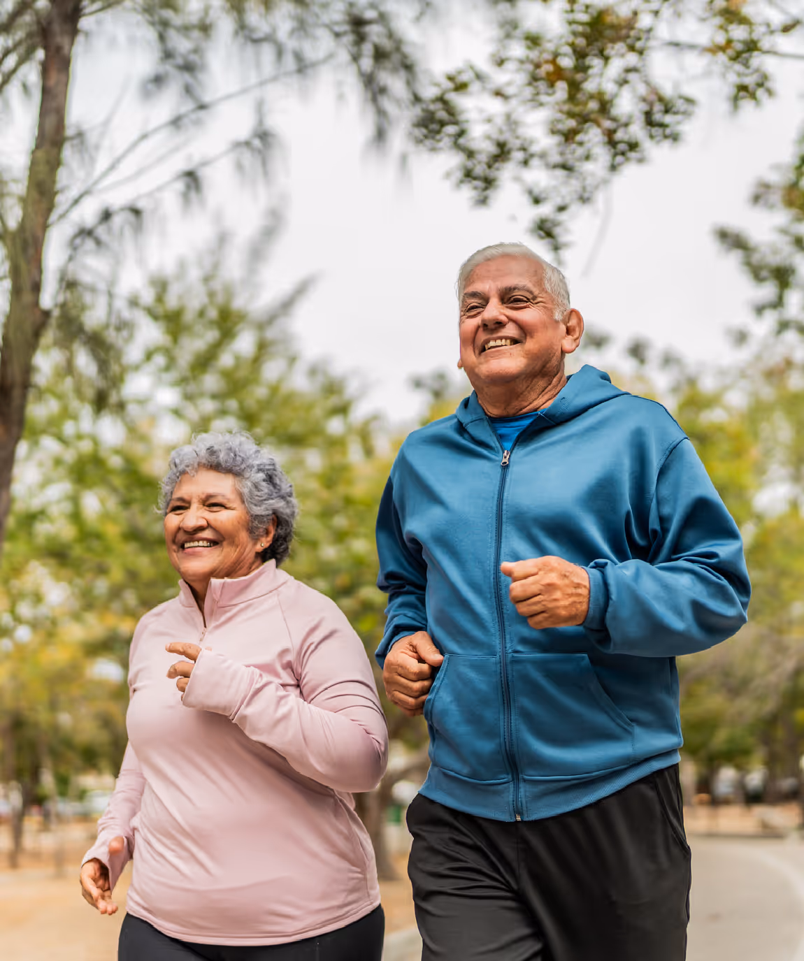 Smiling elderly couple jogging outdoors on a park trail surrounded by trees.