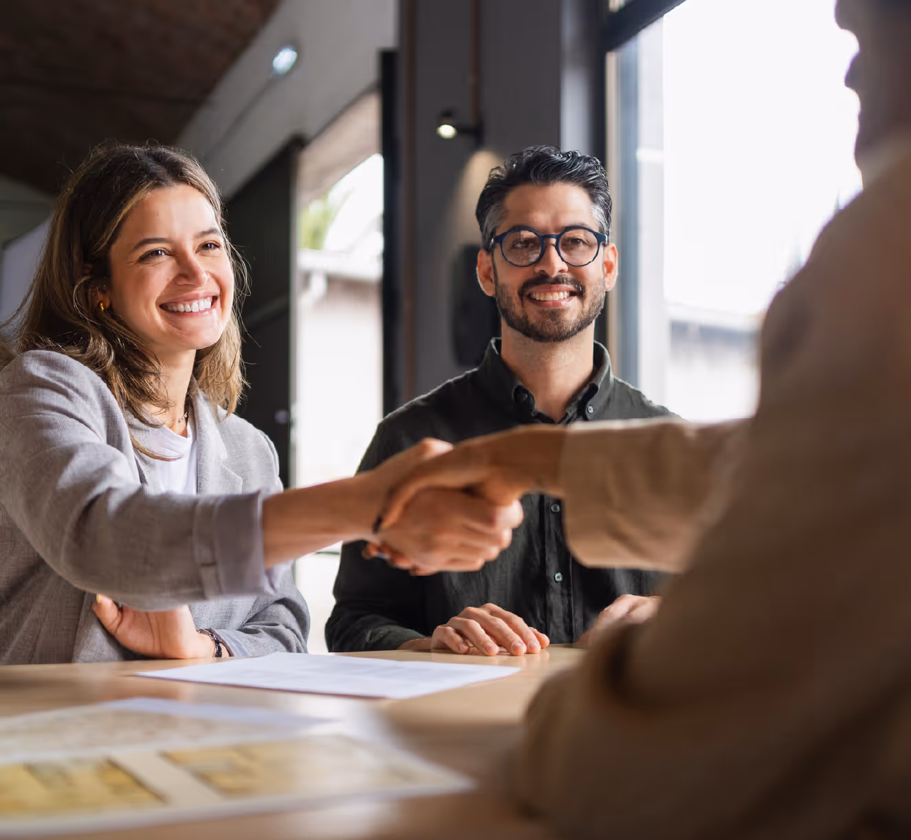 Smiling businesswoman shaking hands with another person while a man with glasses looks on during a meeting.