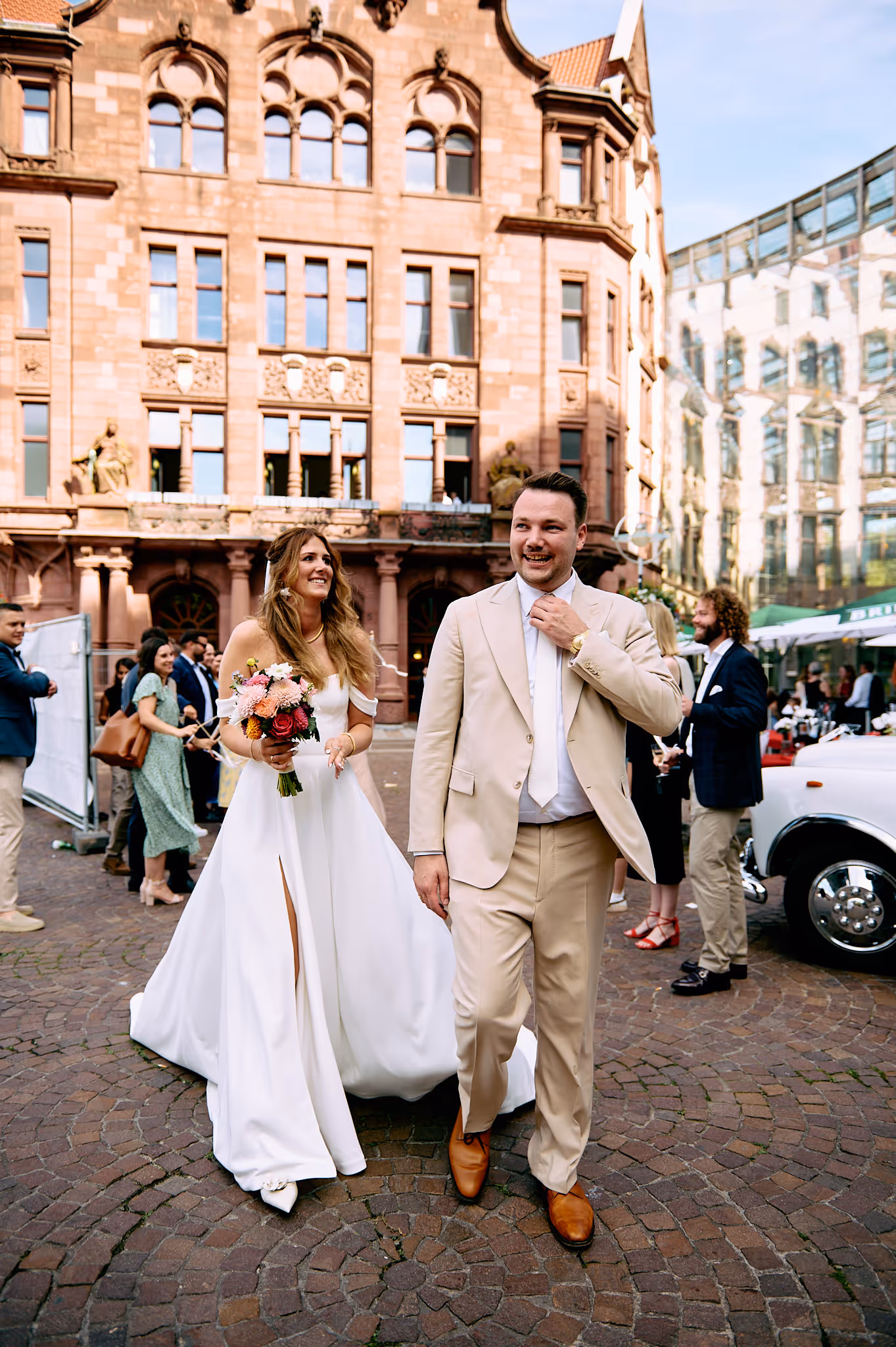 Das frisch verheiratete Hochzeitspaar kommt aus der Kirche, umgeben von seinen Gästen. Der Mann trägt einen Maßanzug, als Einreiher mit passender Hose in einem hellen Beige. Die Sonne scheint und beide lachen glücklich.
