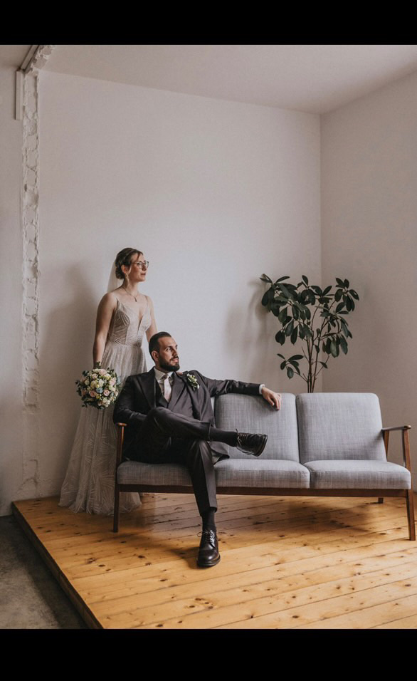Junges Hochzeitspaar, der Mann sitzt auf dem Sofa in einem dunkel grauen dreiteiligem Anzug, bestehend aus einer einreihigen Jacke, einer Weste und einer Hose. Alles aus reiner Schurwolle. Die Frau steht in ihrem langen weißen Hochzeitskleid daneben und hält ihren Brautstrauß aus weißen Blumen.