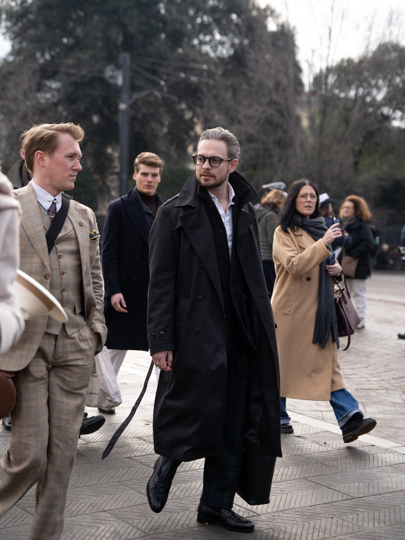 A man walking down the street. He is wearing a long black cotton trench coat with a belt, a black teddy boucle safari jacket, black watch flannel pants and black penny loafer. 