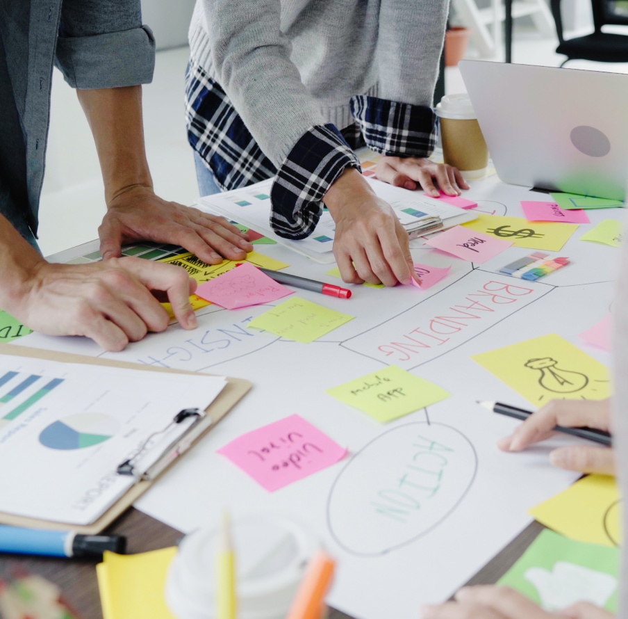 The hands of a group of people gathered around a table covered with colorful sticky notes and paper charts. The hands reaching out to arrange and discuss the materials on the table.