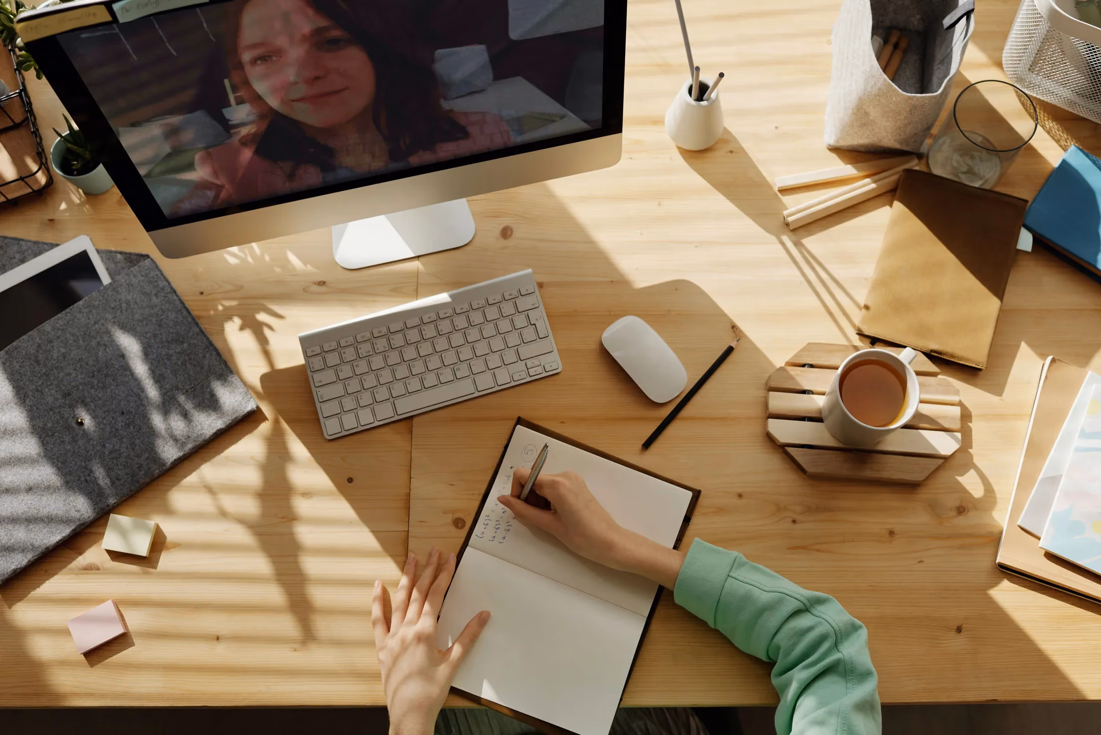 Plan de bureau en bois avec clavier, souris, carnet, tasse de café et une personne écrivant des notes pendant une visioconférence sur un écran d’ordinateur.