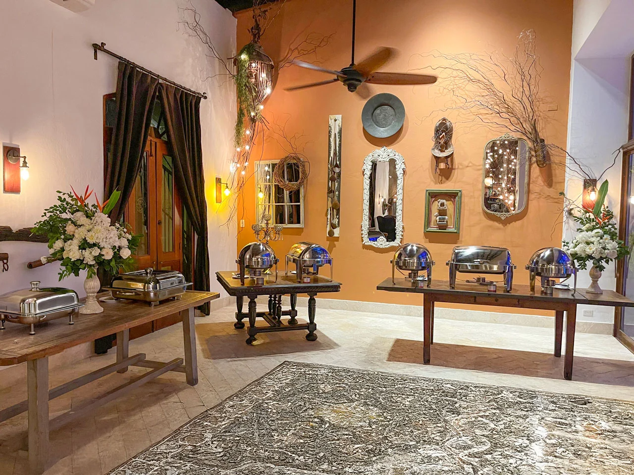 Indoor buffet setup with wooden tables, chafing dishes, floral arrangements, and decorative wall mirrors and ornaments.