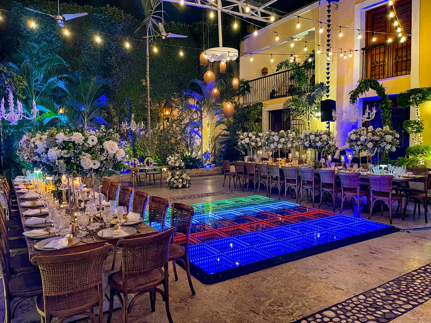 Outdoor nighttime banquet setting with two long decorated tables, floral centerpieces, candles, and a multicolored LED dance floor in the center.