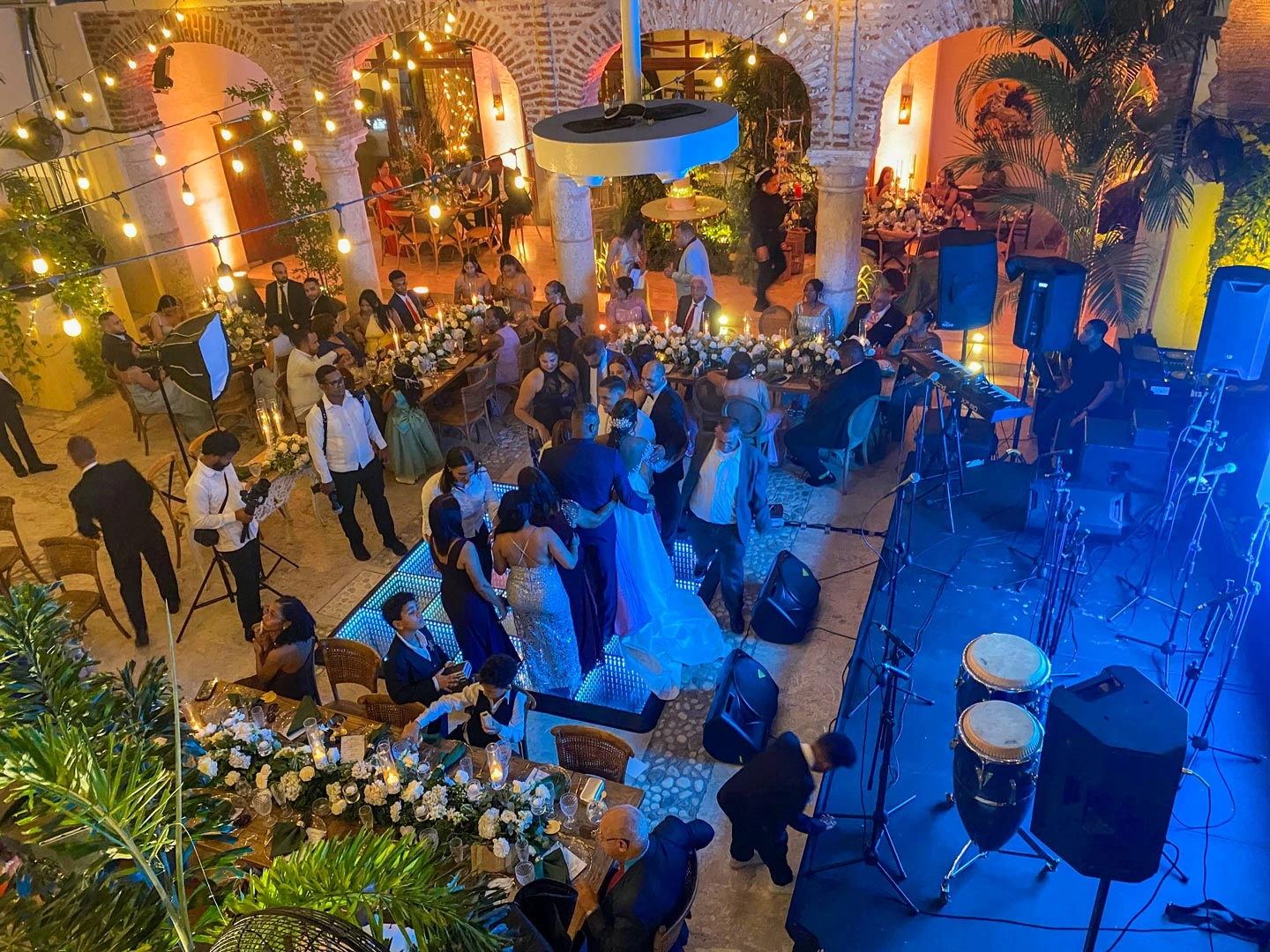 Indoor wedding reception with guests seated at flower-adorned tables and a group dancing on an illuminated dance floor near a stage with musical instruments.