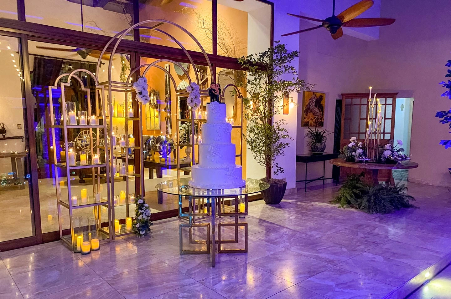 Elegant indoor wedding setup with a four-tier white cake on a glass and gold geometric table, surrounded by candle-lit gold arches and floral arrangements.