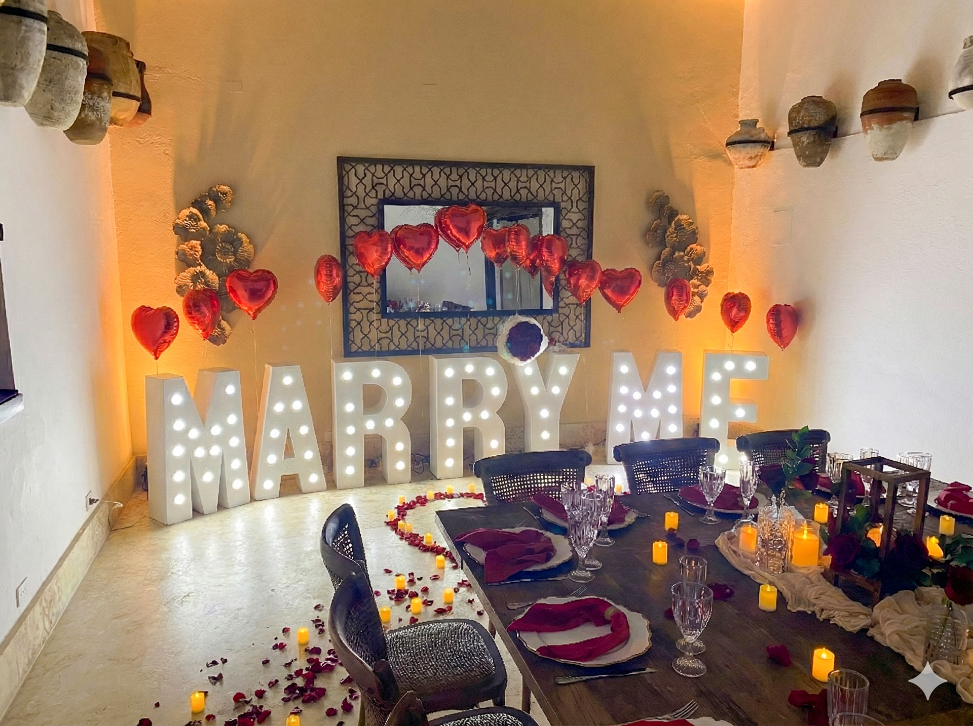 Romantic indoor dining setup with illuminated 'MARRY ME' letters, red heart-shaped balloons, candles, and rose petals.