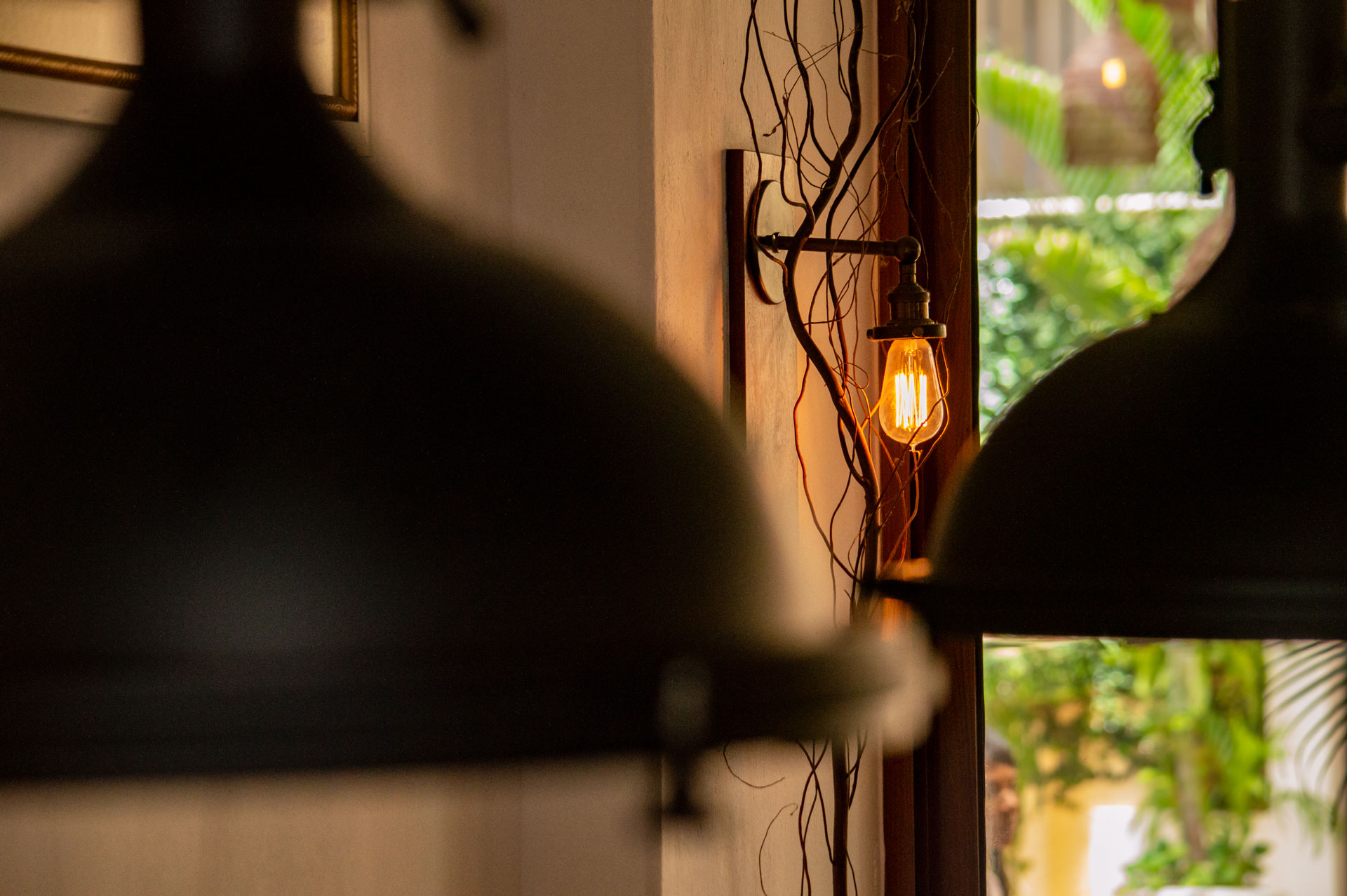 A vintage-style exposed filament light bulb mounted on a wall surrounded by decorative branches, with blurred black hanging lamps and lush greenery outside a window.