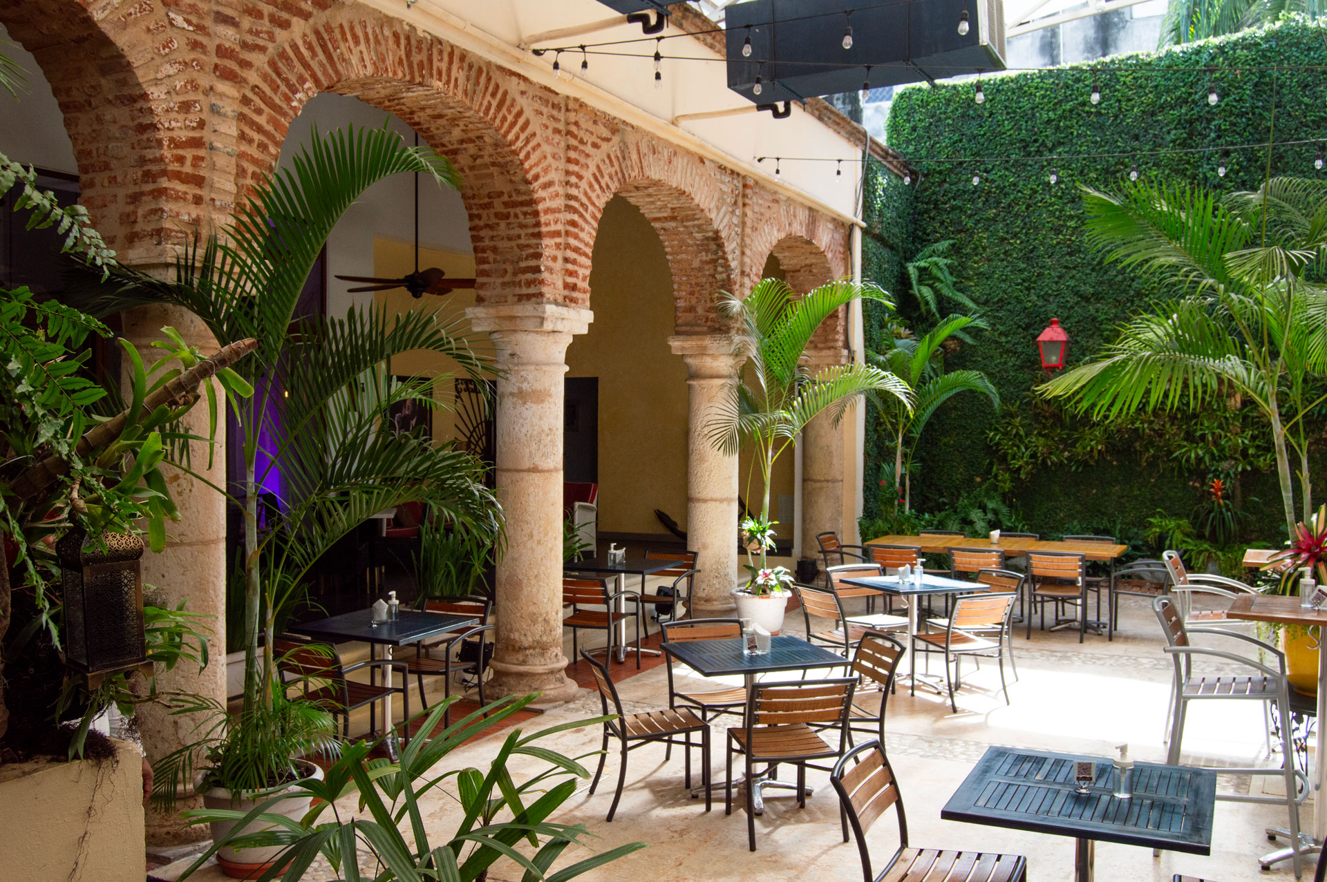 Outdoor courtyard café with stone arches, wooden tables and chairs, surrounded by lush green plants and a tall ivy-covered wall.