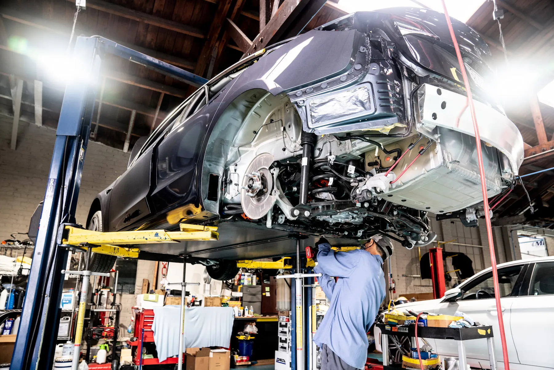 Mechanic working under a car lifted on a hydraulic lift inside an automotive repair shop.